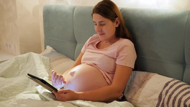 Portrait of smiling pregnant woman in pajamas lying in bed at night and browsing internet or social media on tablet computer