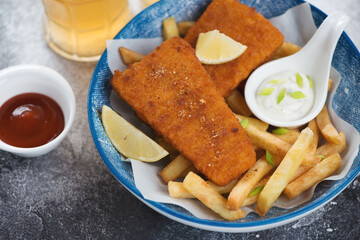 Deep-fried breaded fish fillet with french fries and dips served in a blue bowl, closeup, studio shot