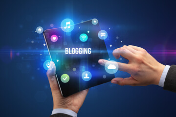 Businessman holding a foldable smartphone with BLOGGING inscription, social media concept