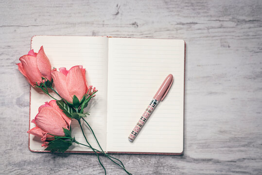 libreta de apuntes en una mesa de madera blanca junto a un lapicero y unas rosas color salm&oacute;n 