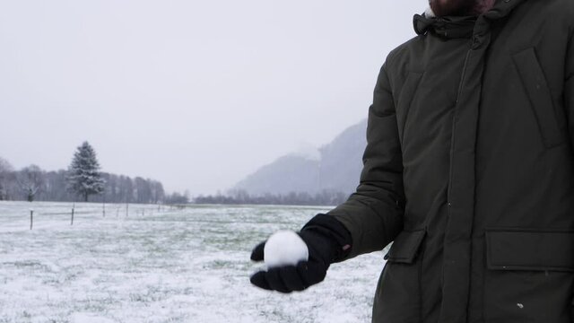 Person playing with snowball outdoors on grass field, making tricks with hand. Slow motion close up with snow covered trees in backdrop.