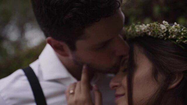 Young happy bride with a wreath in her black hair caress her groom with on her left hand a wedding ring while he is kissing her forehead. Close up shot