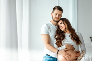 A pregnant girl with her husband are standing in a bright bedroom. Beautiful belly of a young attractive pregnant girl. Family, marriage, childbirth concept.
