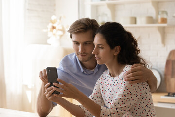 Millennial Caucasian man and woman sit at home kitchen speak on video call or have webcam talk on smartphone. Young couple spouses use cellphone look at screen shopping on internet on gadget.