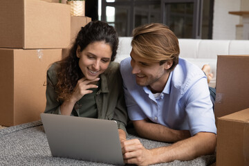 Fototapeta na wymiar Happy millennial Caucasian man and woman lying on floor in new home on relocation day browsing wireless internet on laptop. Smiling young couple renters search for house rent on computer online.