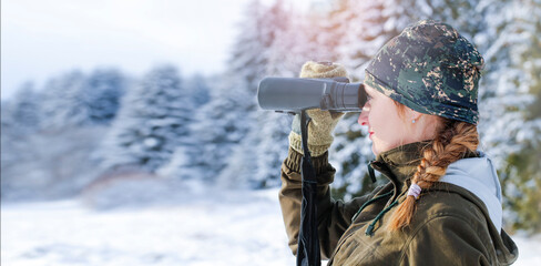 A young hunter woman in the snowy forest. She watches wild animals in the forest using binoculars. Winter landscape.