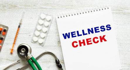 WELLNESS CHECK is written in a notebook on a white table next to pills and a stethoscope.