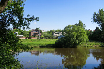 Close-up of cottages on the river bank, with a reflection in the water. Private houses, village on a hill against the blue sky. Lawn with trees and green grass in front of the house.