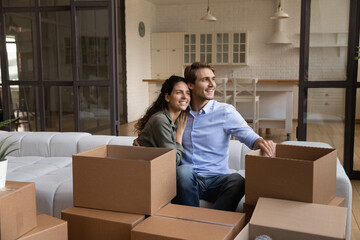 Smiling young Caucasian man and woman sit on sofa hugging look in distance dream together. Happy millennial couple renters enjoy moving relocation day to new own home or house. Rent, estate concept.