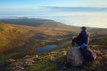 Naklejka na ściany i meble Hiker man sitting on the big stone on the top of the mountain and looking at morning landscape with mountains, lake and cloudy sea. Isle of Skye, Scotland