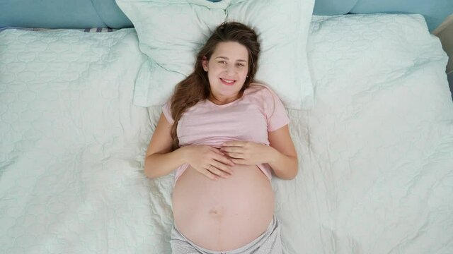 Top view of happy smiling pregnant woman enjoying pregnancy lying on bed and stroking her big belly. Concept of pregnancy, preparing and expecting child.