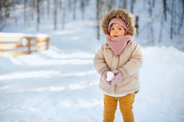 Fototapeta na wymiar Adorable baby girl in a warm snow suit playing in snow on a very sunny and clear winter day in a park.