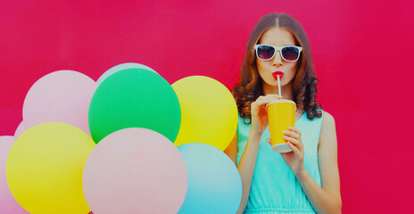 Portrait of young woman drinking a juice with colorful balloons on a pink background