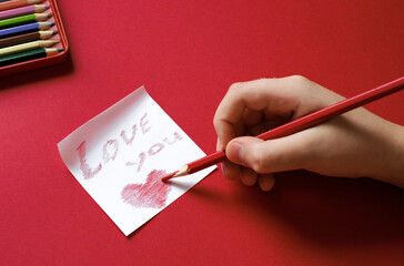 Hands paint heart and love you on white sticker in handwriting with pencil on a red background.