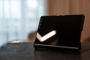 A black tablet stands on a stone table against a blurred background of the room. Copy space.
