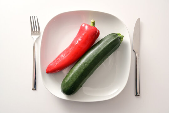 Raw red pointed pepper and zucchini on a white plate and cutlery on a bright background, healthy diet with mediterranean vegetables to lose weight, copy space, high angle view from above © Maren Winter