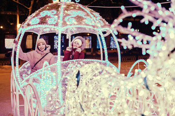 Girl friends having fun in illuminated fairy tale carriage with horses decorated with lights and garlands for Christmas night and New Year holiday. Friendship and winter vacation concept