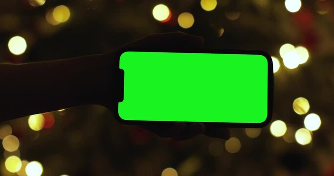 Close up hand holding iPhone 12 Pro Max with a horizontal landscape green screen at home. Background evening lights garlands Christmas. Smartphone technology cell phone message.