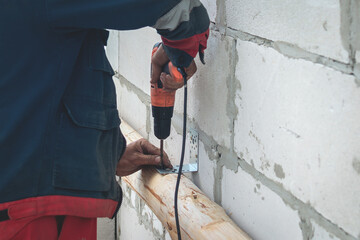 A male worker in overalls uses an electric screwdriver to screw screws into a wooden beam. Construction, installation, fixing of structures outdoors
