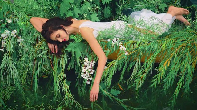 Fantasy happy brunette woman lies in boat, gently touches a water her hand. Girl princess in white long dress floats on lake. Friday, relaxation and enjoyment. Creative wedding decor. Nature greens 