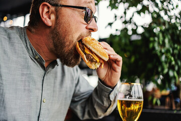 Middle aged bearded hungry man sitting in restaurant and eating delicious burger.
