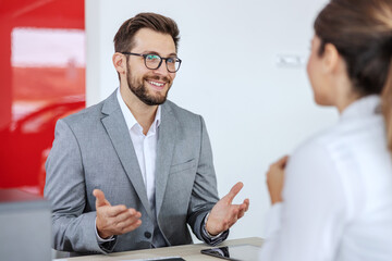 Smiling friendly car seller sitting in car salon with a woman who want to buy a car and making an...