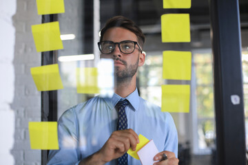 Young modern business man using adhesive notes while standing behind the glass wall in the office.