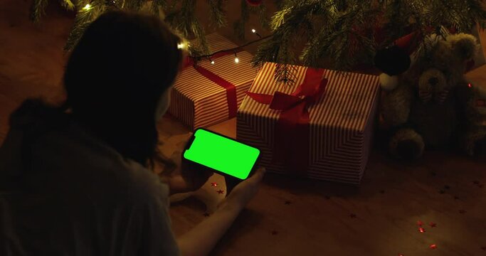 Middle shot of woman lie on floor, texting, typing, chatting on phone next to christmas present, smiling in living room. Concept of family holidays. iPhone 12 green screen.