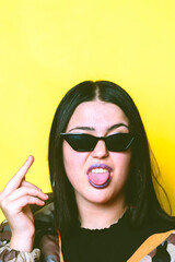 Teenager with sunglasses and yellow background sticking out his tongue and reading the middle finger.