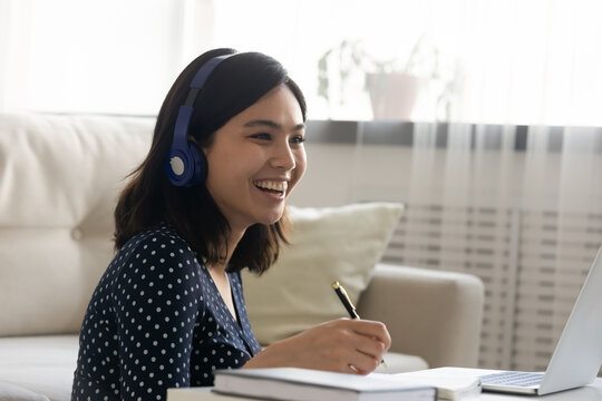 Happy Millennial Vietnamese Asian Woman Wearing Wireless Headphones, Enjoying Funny Educational Webinar On Computer, Watching Video Lecture Or Involved In Discussing Material Online At Home.