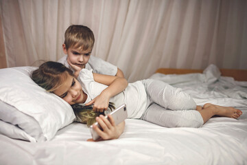 Two siblings watching video in their mobile electronic device in the bed, gadget dependence and addiction, online and internet for the whole day, family lifestyle, indoor