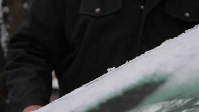 Person hand moves black scraper on car windshield and small ice pieces fall on hood slow motion