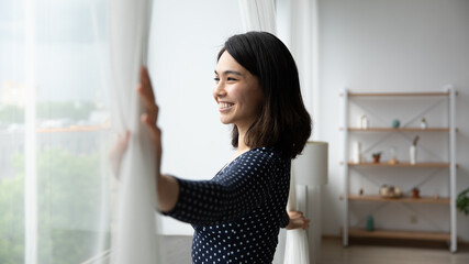 Side view happy millennial asian korean woman opening curtains, admiring morning cityscape view, smiling beautiful lady enjoying peaceful morning time, woke up early on weekend starting new day.