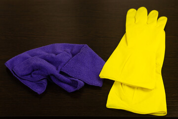 Yellow rubber protective gloves and a purple rag, microfiber, lie on a dark wooden background. General or regular cleaning. Commercial cleaning company. Cleaning items.