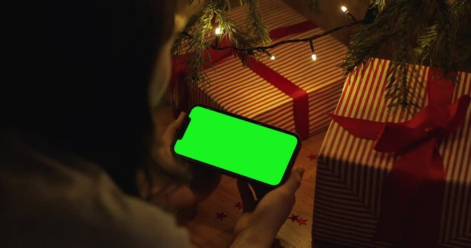 Over the shoulder hand holding iPhone 12 Pro Max with a horizontal landscape green screen at home. Background evening lights garlands Christmas