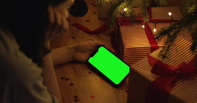 Girl hand holding the smartphone with green screen on Christmas decorated background. Close-up woman watching video on iPhone 12 Pro Max staying at home.