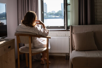 Back view of beautiful young woman in bathrobe sitting on armchair and enjoying near open window at sunny morning.