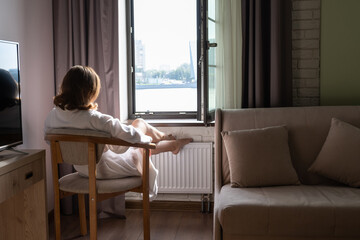 Back view of beautiful young woman in bathrobe sitting on armchair and enjoying near open window at sunny morning.