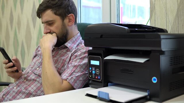 The printer on the table printed the word smile at the man next to it. Man in the office uses a smartphone at the workplace
