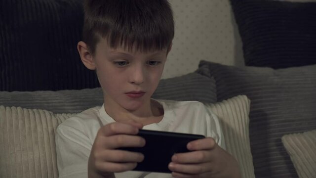 White boy 7-9 years old enthusiastically playing a game on a smartphone, sitting on the couch. Children's dependence on gadgets.