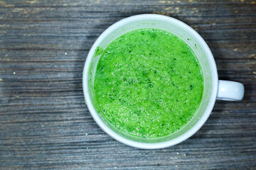 Top view to white round wide cup with fresh green soup. Aged wooden grey background. Raw vegetarian food. Vegetable smoothie. Vitamins and healthy dieting. Seasonal spring cocktail. 