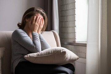 Fototapeta premium Sad scared woman covering her face with hands and crying. Lonely girl sitting near window with pillow on a sofa of apartments.