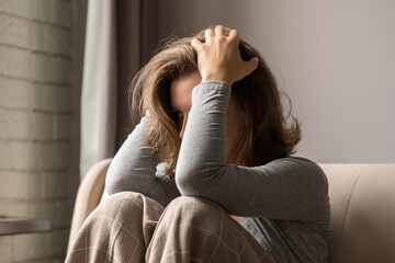 Obraz premium Sad scared woman in panic attack grabbing her head by hand. Lonely girl wrapped in a plaid sitting near window of apartments.