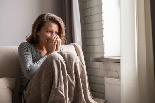 Sick Brunette Woman Sneezing Sitting On The Couch In The Living Room Near Window