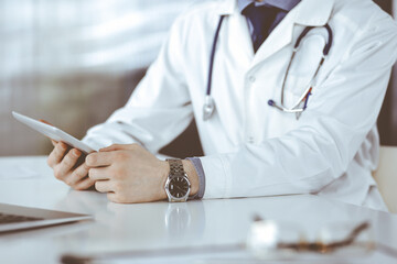 Unknown male doctor sitting and working with tablet computer in clinic at his working place, close-up. Young physician at work. Perfect medical service, medicine concept