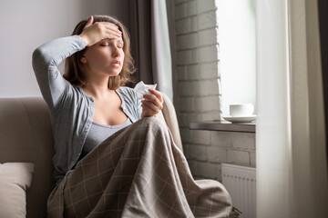 Sick brunette woman with headache having cold and flu sneezing sitting on the couch in the living room near window