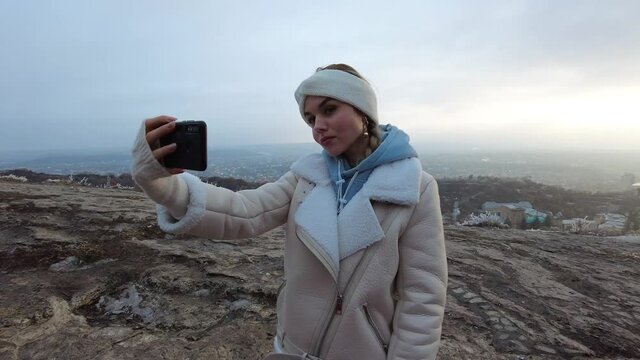 A young girl in the winter season makes a selfie on the phone in a tourist place on Mount Mashuk, in the background you can see the city and trees in hoarfrost