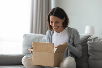 Close up overjoyed smiling young woman opening cardboard box with awaited parcel, sitting on couch...