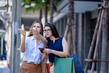 Two young women with shopping bags and pointing on direction and checking way on smartphone in city,Sale,Consumerism and People Concept.