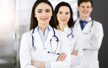 Fototapeta premium Group of young professional doctors standing as a team with arms crossed in sunny hospital. Medical help, insurance in health care, best disease treatment and medicine concept